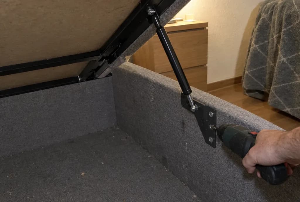 Mano con un atornillador soltando el soporte metálico del amortiguador de gas en el lateral de un canapé abatible abierto y vacío