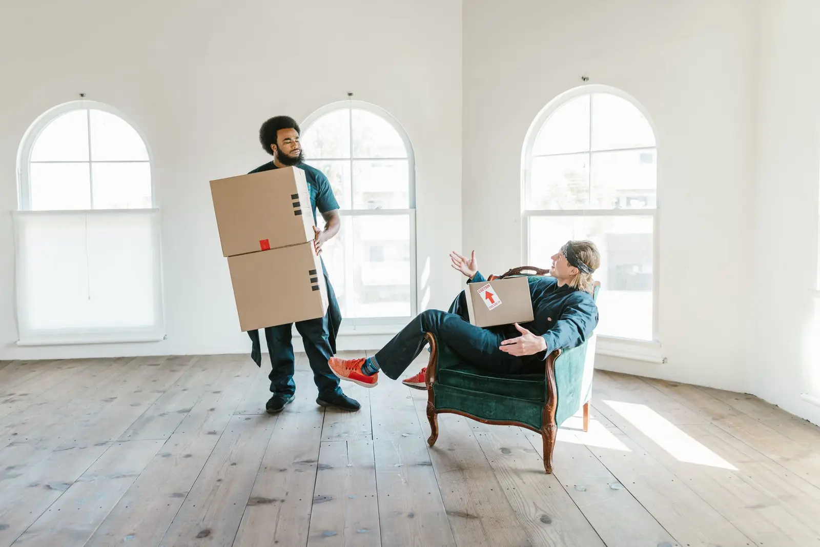Habitación vacía con ventanales en arco y suelo de madera, con dos personas y cajas de cartón